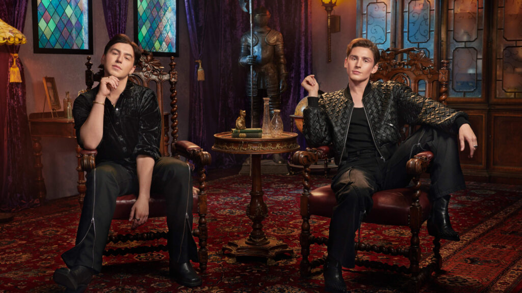 Two young men seated in ornate wooden chairs in a vintage room with stained glass windows and a suit of armor.