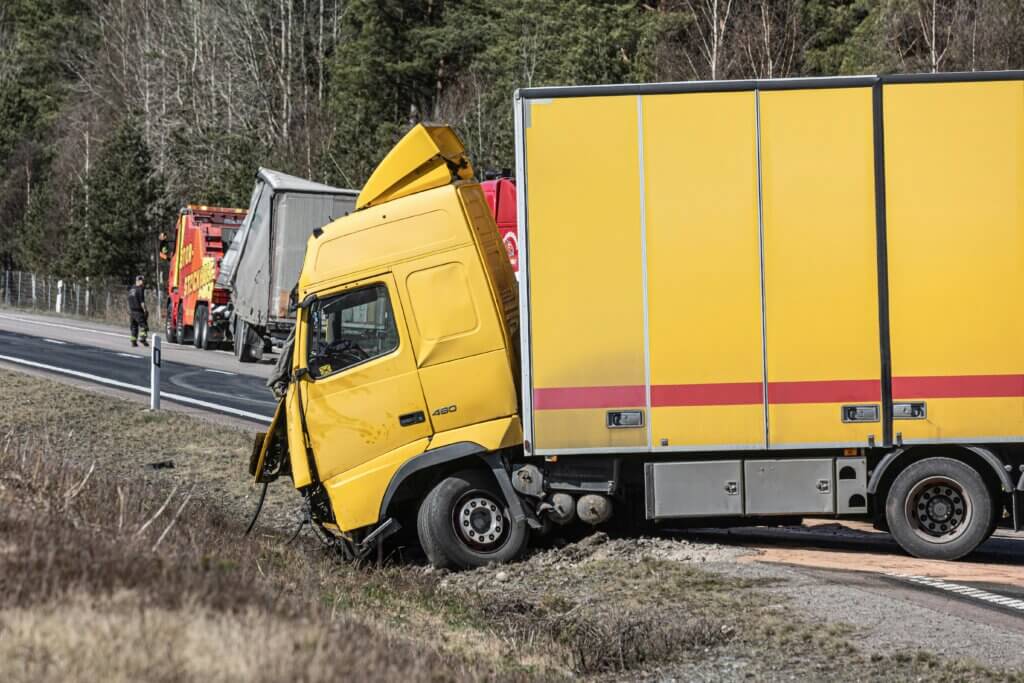 Yellow semi-truck with front-end damage stuck off the road with a red tow truck and worker in the background