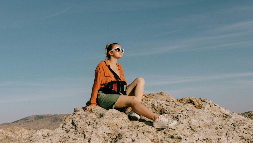 Woman wearing white sunglasses and an orange shirt sitting on a rocky hill with a Kenzo bag under a clear sky