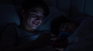 Teen boy smiling while using a smartphone in bed next to a sleeping person in a dark room