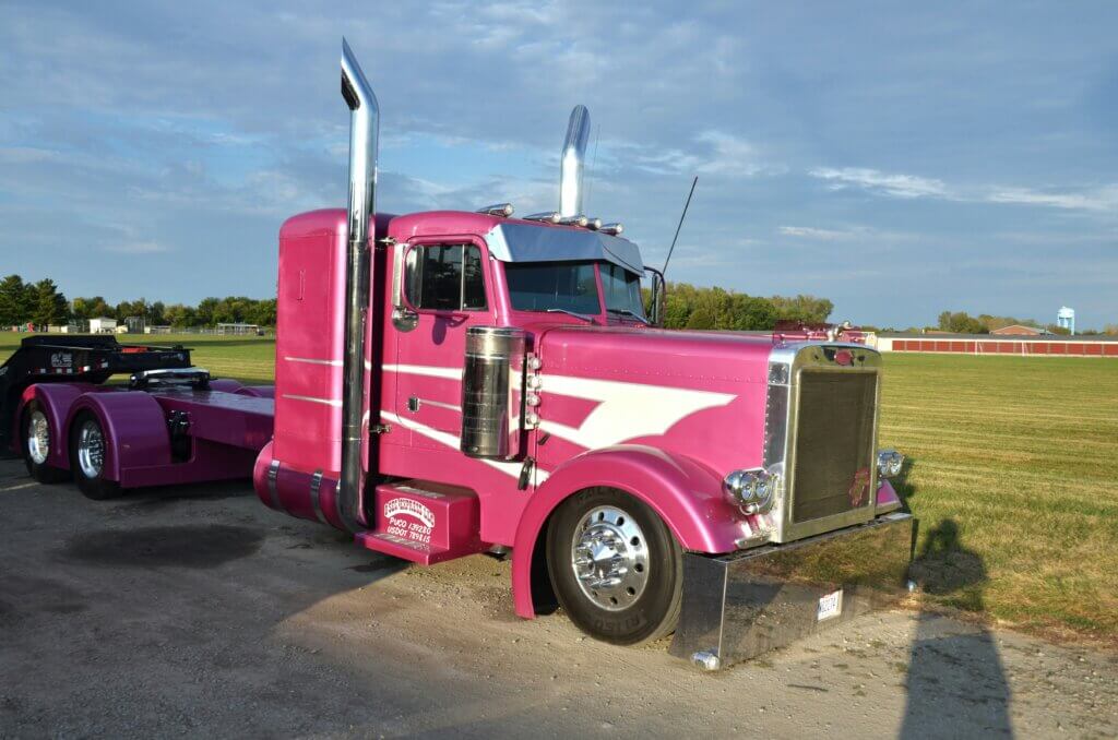 Pink Peterbilt semi-truck with white stripe parked on gravel near a grassy field under a partly cloudy sky.