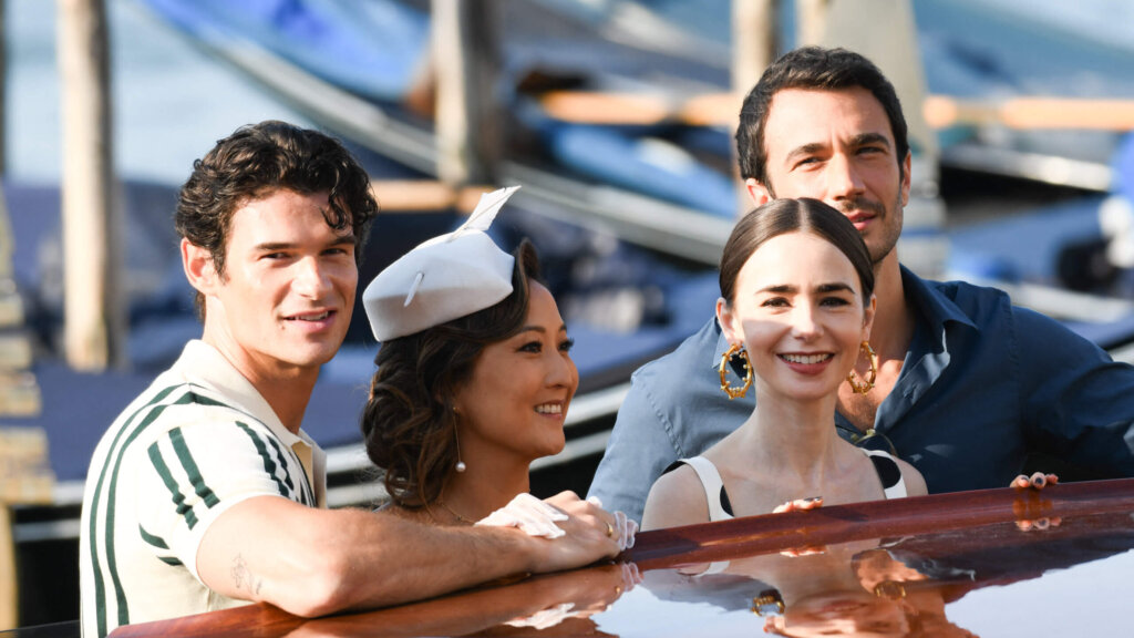 Four young adults smiling and leaning on a wooden boat near a dock with gondolas in the background