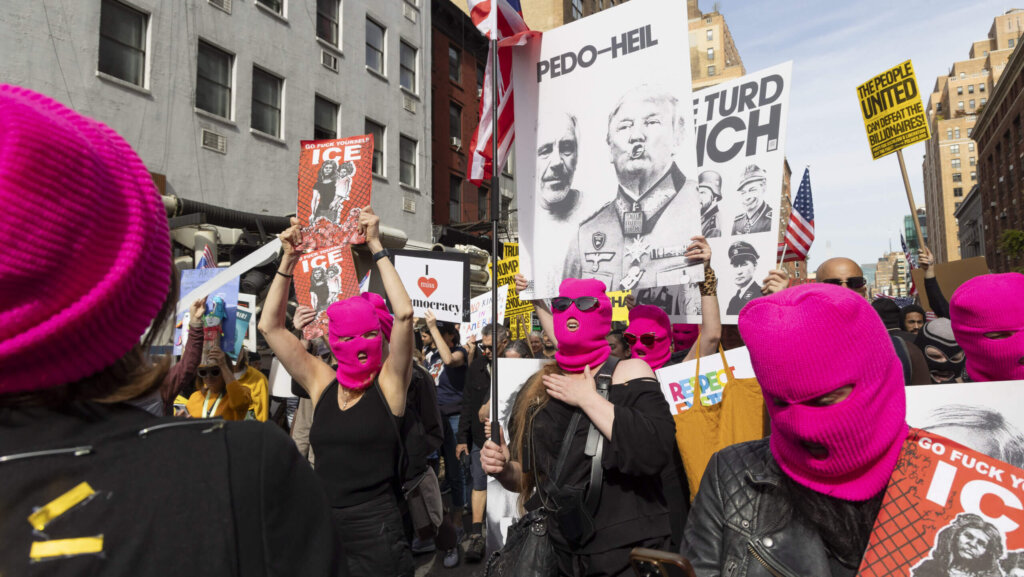 Protesters wearing pink ski masks hold signs criticizing Donald Trump and ICE during a street demonstration.