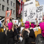 Protesters wearing pink ski masks hold signs criticizing Donald Trump and ICE during a street demonstration.