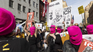 Protesters wearing pink ski masks hold signs criticizing Donald Trump and ICE during a street demonstration.