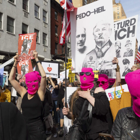 Protesters wearing pink ski masks hold signs criticizing Donald Trump and ICE during a street demonstration.