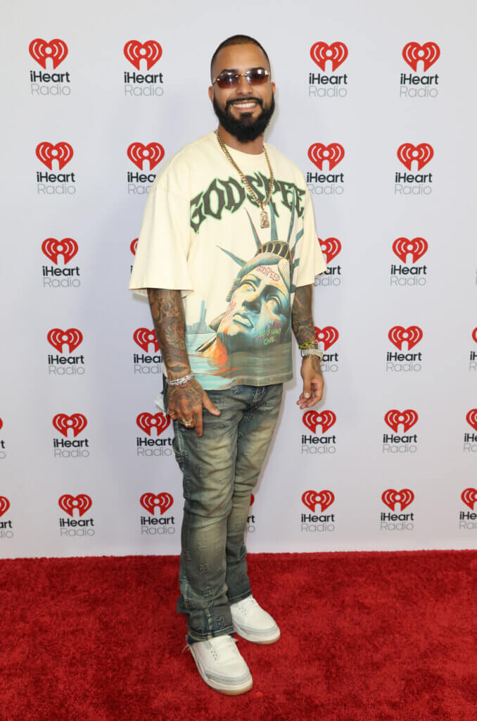 Man wearing sunglasses, a Godspeed Statue of Liberty graphic t-shirt, jeans, and white sneakers on an iHeartRadio red carpet