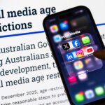Hand holding smartphone displaying social media apps in front of text about Australian social media age restrictions.