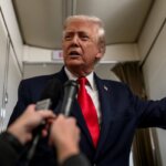 Donald Trump speaking to reporters inside an airplane cabin, wearing a navy suit and red tie.