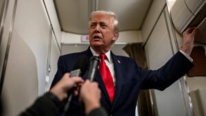 Donald Trump speaking to reporters inside an airplane cabin, wearing a navy suit and red tie.