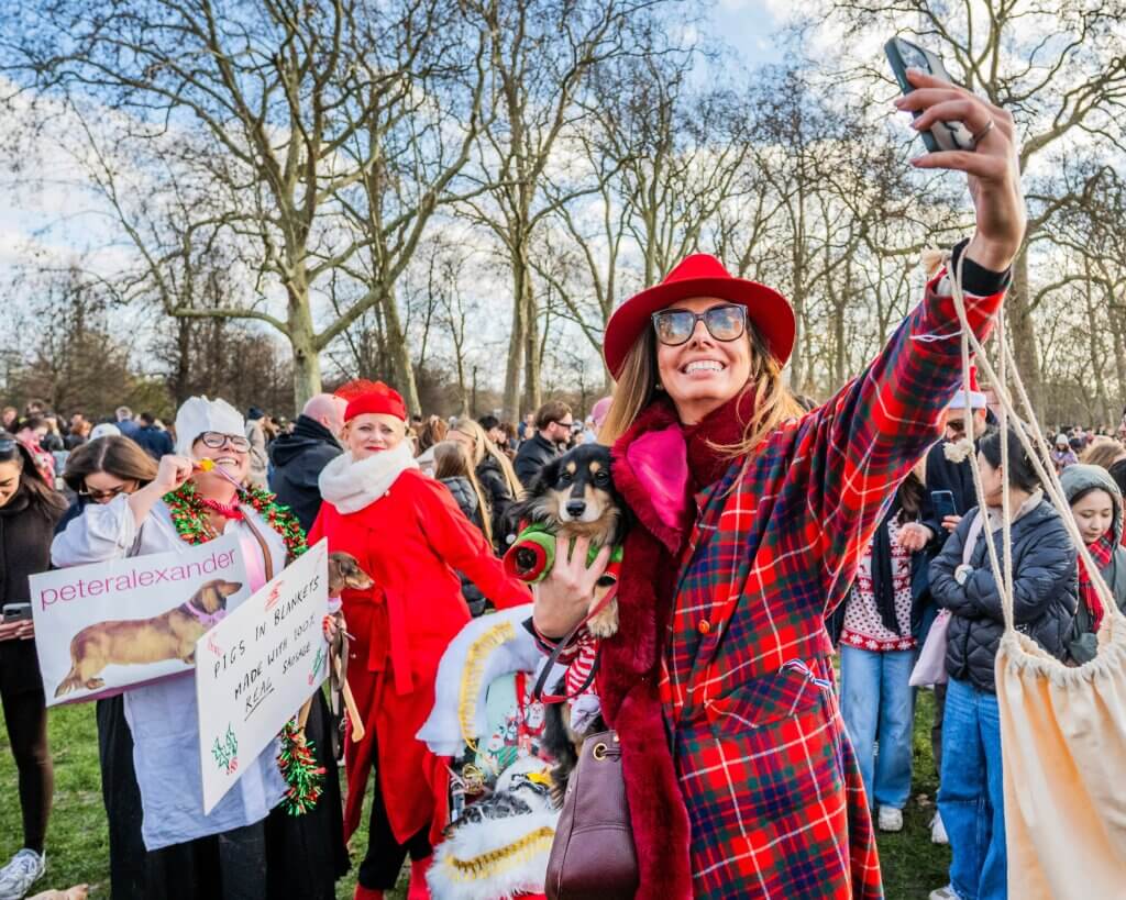 Woman in red plaid coat and hat taking a selfie while holding a dog at a festive outdoor gathering with people in holiday attire.