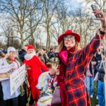 Woman in red plaid coat and hat taking a selfie while holding a dog at a festive outdoor gathering with people in holiday attire.