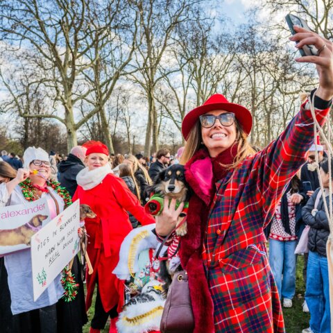 Woman in red plaid coat and hat taking a selfie while holding a dog at a festive outdoor gathering with people in holiday attire.