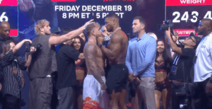 Jake Paul and Tommy Fury face off during a weigh-in event with photographers and spectators around them.