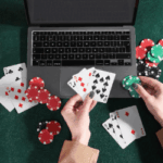 Hands holding playing cards and poker chips in front of a laptop on a green felt surface.