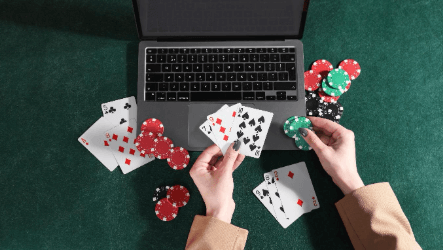 Hands holding playing cards and poker chips in front of a laptop on a green felt surface.