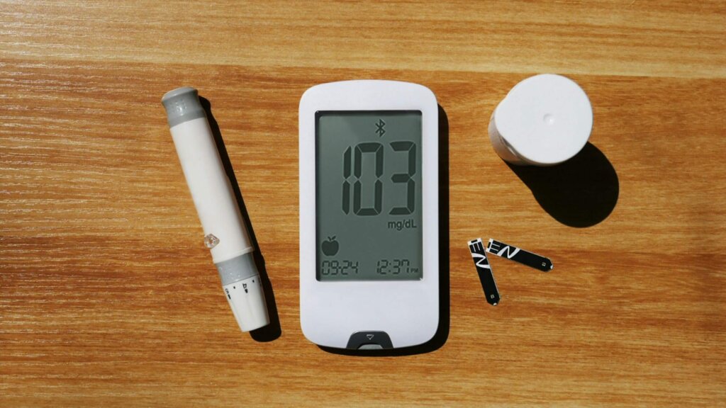 Blood glucose meter displaying 103 mg/dL with lancet device, test strips, and container on wooden surface.