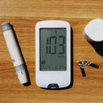 Blood glucose meter displaying 103 mg/dL with lancet device, test strips, and container on wooden surface.