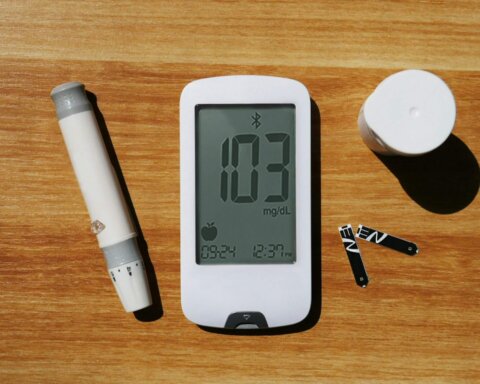 Blood glucose meter displaying 103 mg/dL with lancet device, test strips, and container on wooden surface.