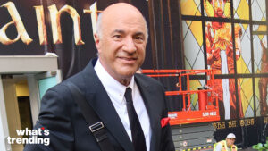 Kevin O'Leary in a black suit and tie with a red pocket square smiling outdoors near a stained glass backdrop.