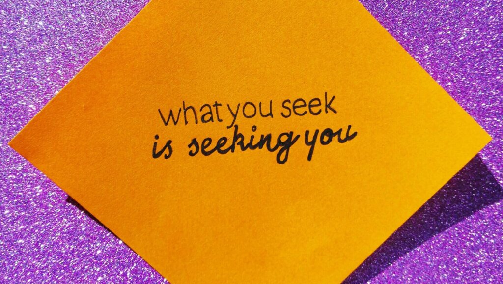 Orange sticky note with the handwritten quote "what you seek is seeking you" on a purple glitter background