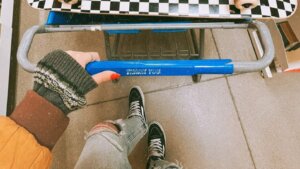 Person pushing a shopping cart with a checkerboard skateboard, wicker basket, and clothes inside a store aisle.