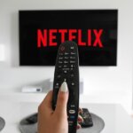 Hand holding TV remote pointed at screen displaying Netflix logo in modern living room.