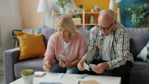 Elderly couple sitting on a couch reviewing bills and counting cash at a white table in a living room.
