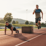 Man doing push-ups and another man jumping over a wooden box on an outdoor running track at sunset