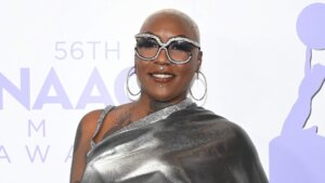Person with short blonde hair wearing large rhinestone sunglasses and a shiny silver one-shoulder top at the 56th NAACP Awards