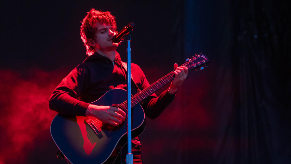 Male musician playing a Gibson acoustic guitar and singing into a microphone on stage with red lighting.