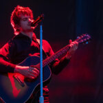 Male musician playing a Gibson acoustic guitar and singing into a microphone on stage with red lighting.