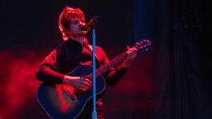 Male musician playing a Gibson acoustic guitar and singing into a microphone on stage with red lighting.
