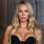 Blonde woman with blue eyes wearing a black strapless dress against a blurred architectural background