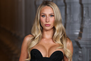 Blonde woman with blue eyes wearing a black strapless dress against a blurred architectural background