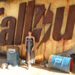 Woman in black dress standing in front of large rusty Fallout sign with blue barrels labeled Prime nearby