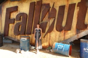 Woman in black dress standing in front of large rusty Fallout sign with blue barrels labeled Prime nearby