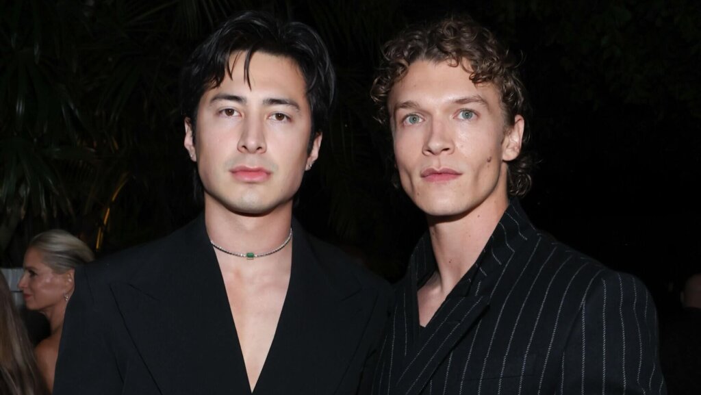Two men wearing black suits, one with a pinstripe pattern and a wristwatch, posing at a nighttime event.