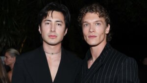 Two men wearing black suits, one with a pinstripe pattern and a wristwatch, posing at a nighttime event.