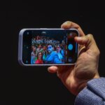 Hand holding smartphone capturing a selfie of a group of people with a large audience in the background.