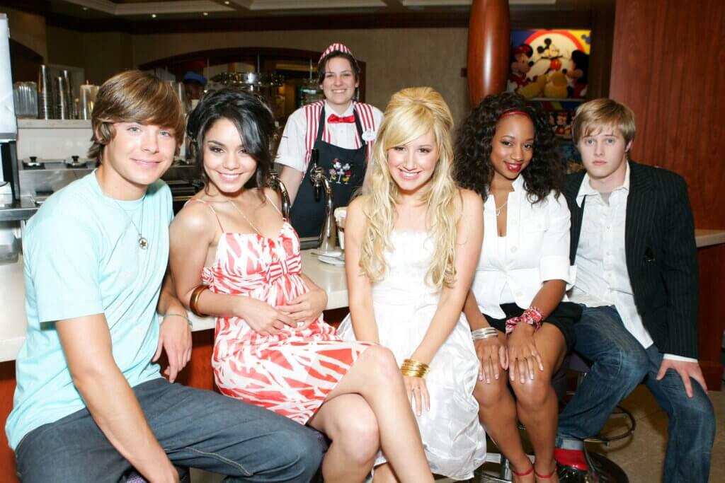 Zac Efron, Vanessa Hudgens, Ashley Tisdale, Monique Coleman, and Lucas Grabeel sitting at a diner counter with a waitress in a Disney-themed uniform behind them.