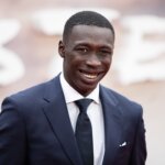 Actor Mamoudou Athie smiling in a navy suit and black tie at a red carpet event