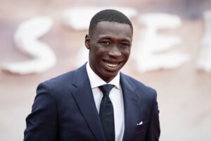 Actor Mamoudou Athie smiling in a navy suit and black tie at a red carpet event