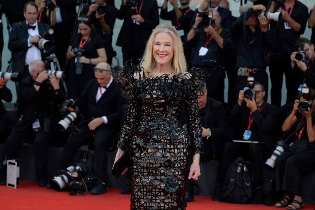 Blonde woman in a black intricate floral dress smiling on a red carpet with photographers in the background