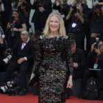 Blonde woman in a black intricate floral dress smiling on a red carpet with photographers in the background