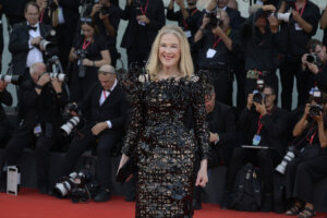 Blonde woman in a black intricate floral dress smiling on a red carpet with photographers in the background
