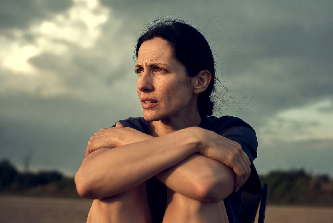 Woman with dark hair sitting outdoors hugging her knees, looking thoughtfully into the distance under a cloudy sky.