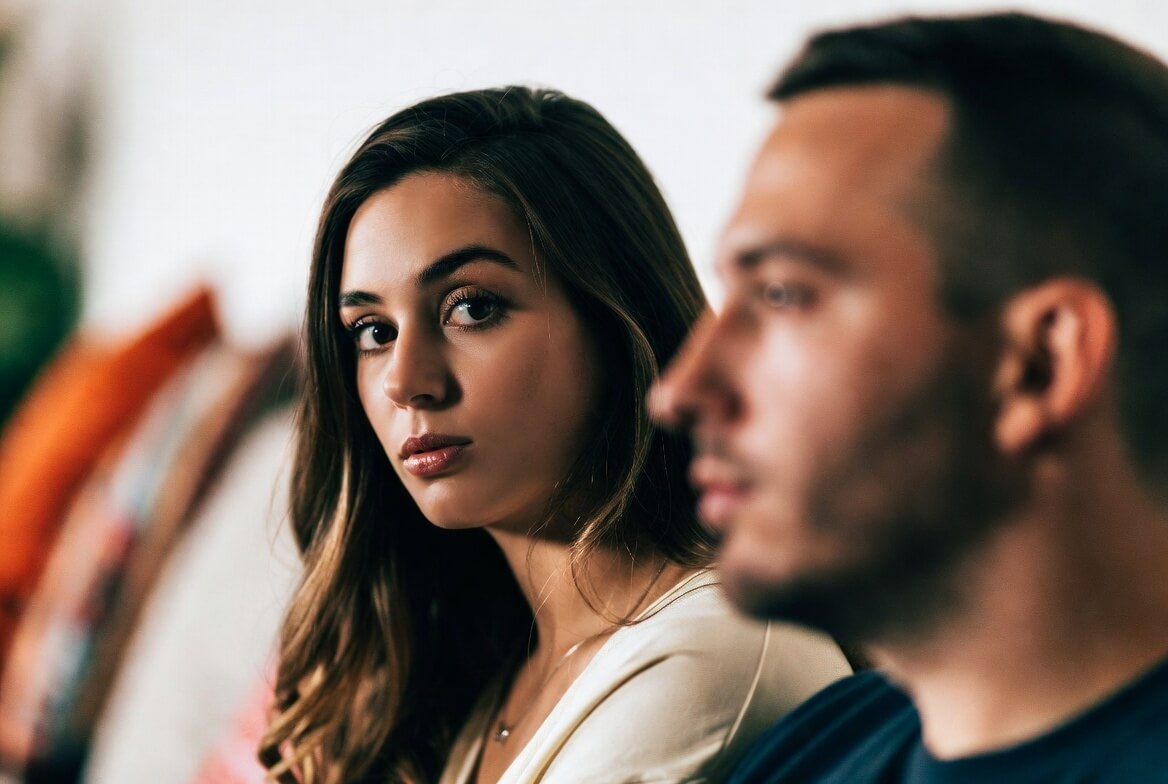 Young woman looking at camera with serious expression, man blurred in foreground facing sideways