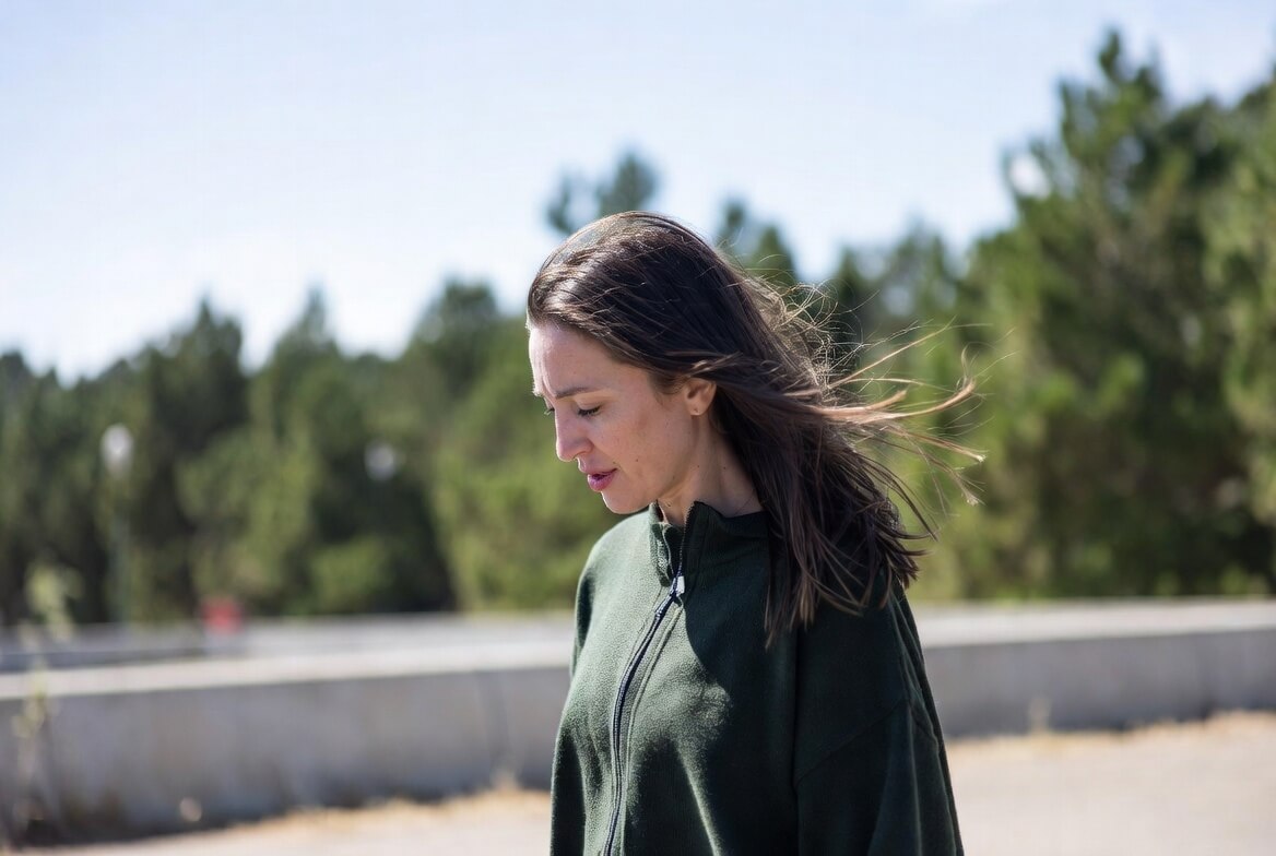 Woman with long brown hair wearing a dark green zip-up jacket outdoors with trees in the background