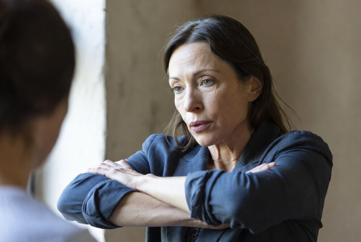 Concerned woman with crossed arms talking to another person indoors near a window
